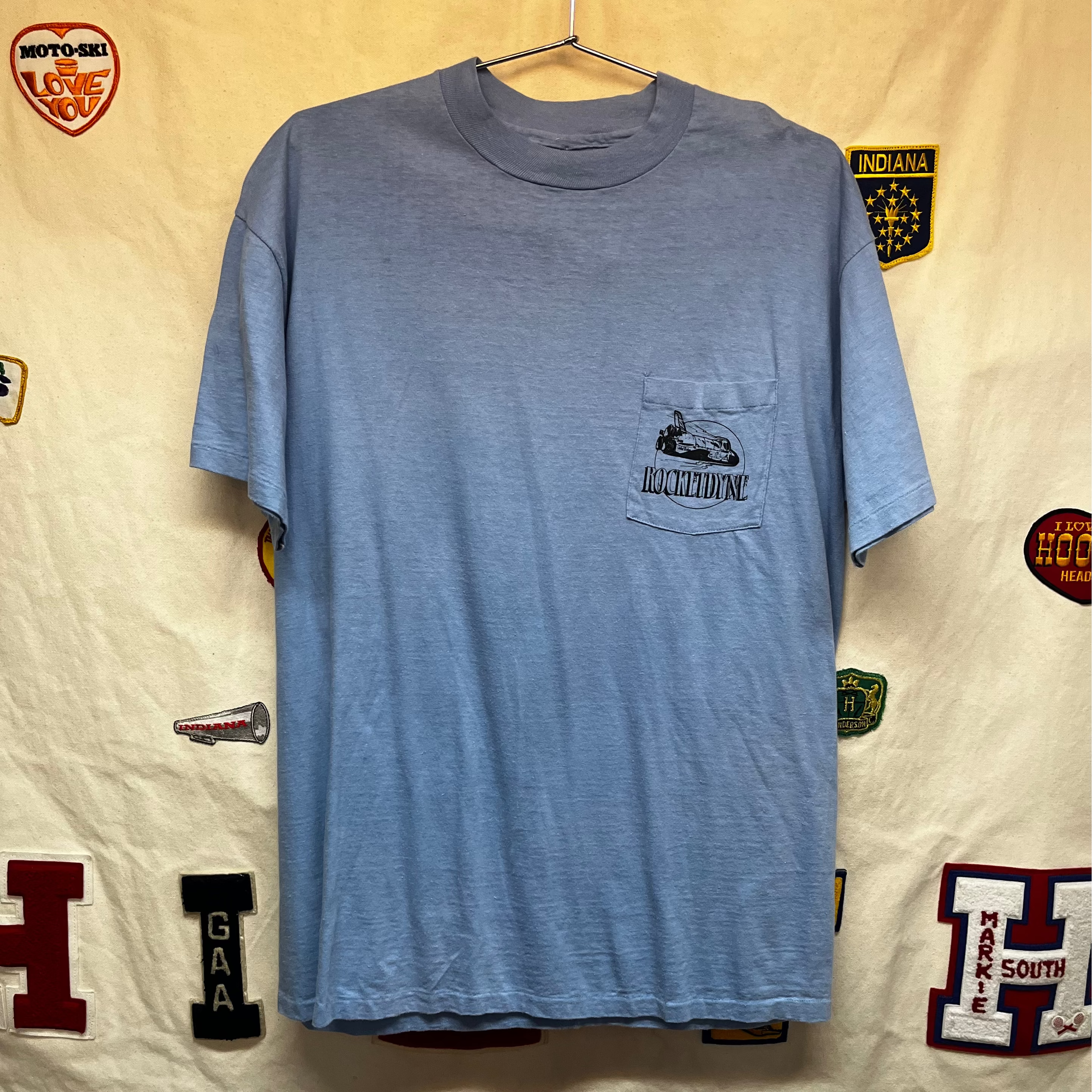 A light blue short-sleeve pocket t-shirt hanging on a hanger against a cream-colored backdrop covered with various vintage patches and insignias. The pocket features a black graphic of a rocket ship and the text 'Rocketdyne"